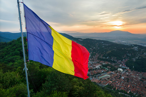 National flag located on the top of the hill with green trees. View of Brasov with multiple buildings on the background at sunset, Romania