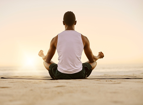 Fit and active man doing yoga, meditating and relaxation exercise on the beach. Calm, peaceful and relaxed male doing breathing exercises and practicing mindfulness by the seashore during sunrise