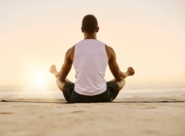 fit and active man doing yoga, meditating and relaxation exercise on the beach. calm, peaceful and relaxed male doing breathing exercises and practicing mindfulness by the seashore during sunrise - men beach back rear view imagens e fotografias de stock