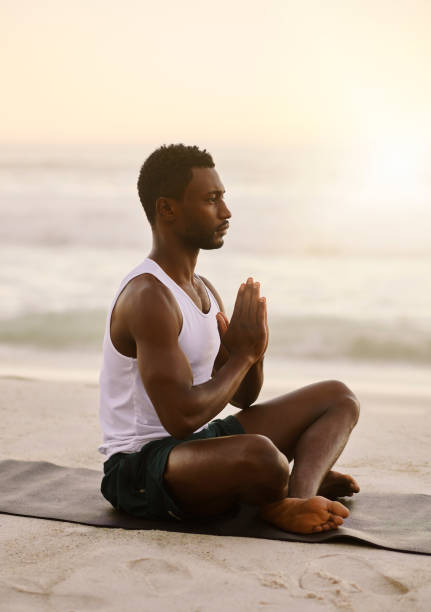 Fit and active man doing yoga, meditating and peaceful exercise on the beach alone. One calm, serious and relaxed black male praying and practicing mindfulness by the seashore during sunrise