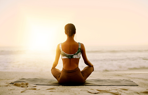 Calm and relaxed woman doing lotus yoga pose exercise and mediation with sunset beach background and copy space. Back view of a fit and healthy yogi woman on a mat practicing inner peace by the ocean