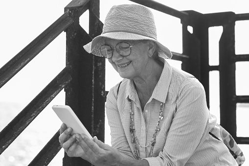 Black and white portrait of smiling senior woman with hat sitting outdoor at sea using mobilel phone
