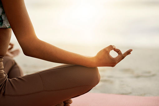 Mindful, calm and relaxed woman meditating outdoors at the beach sitting in the lotus position. Closeup of a Fit and zen female doing yoga during sunrise outside at the sea or ocean shore