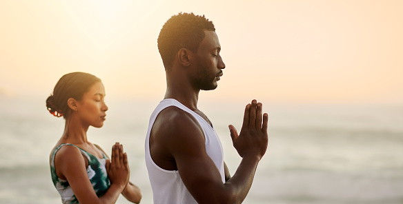 Relaxed, healthy and zen couple meditating on a beach in namaste prayer hands pose by sea and ocean. Calm and spiritual yogis breathing, worshipping and showing thanks in holistic mental health yoga