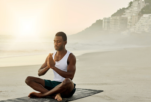 Calm and relaxed African American male doing yoga, meditating on the beach while sitting on a mat. Relaxing, spiritual and peaceful young man in a lotus position with hands together.