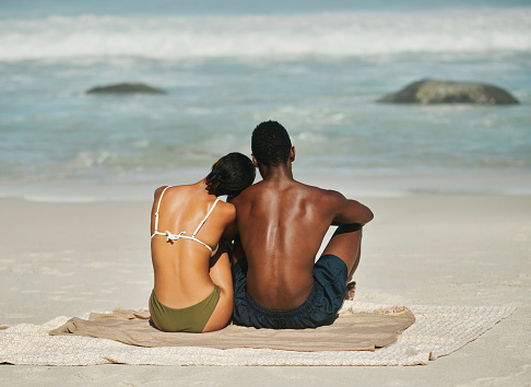 Rear view of young couple sitting and enjoying beach views together. Black boyfriend and girlfriend bonding, showing affection and talking. Sweet lovers relaxing and enjoying free time on the weekend