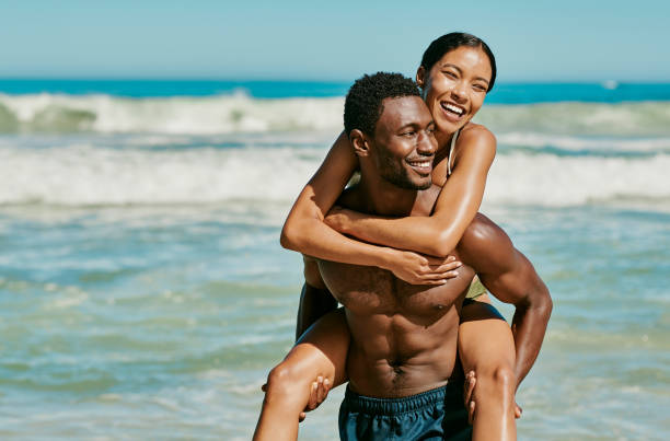 pareja feliz, emocionada y tonta divirtiéndose en verano en la playa en un día caluroso y soleado. hombre afroamericano dando a una hembra un paseo en la orilla del mar junto al océano, riendo y sonriendo de alegría. - sexual activity couple sensuality heat fotografías e imágenes de stock