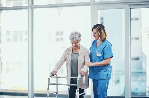 Caregiver helping senior woman walking with a walker in a retirement home showing care and support. Mature or old disabled female patient being assisted by a nurse around the hospital corridor