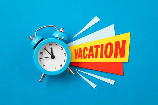 Red alarm clock with colored papers with vacation text on blue background