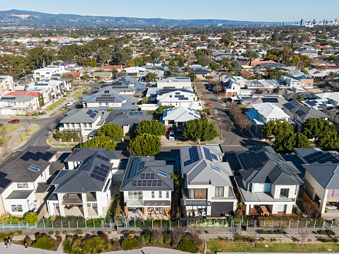 Aerial view looking down on modern housing development with double-storey (two-level) dwellings, landscaped gardens, and established suburb in background with city and hills in distance: solar panels on rooftops, public space