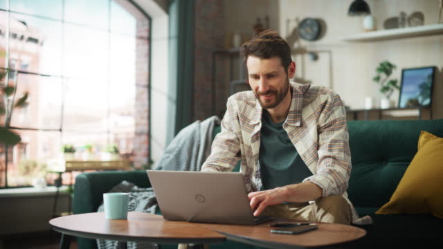Portrait of Smiling Middle Aged Man Working from Home on a Laptop Computer in Sunny Cozy Apartment. Successful Male Entrepreneur Does Remote for e-Business Project, Online Shopping. Zoom Out