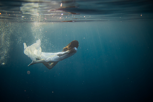 Woman with white dress is diving in the sea, lots of air bubbles surround her after she jumped in the water, view from underwater