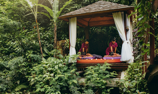 People having massage at spa resort surrounded by trees. Outdoor spa center at luxury holiday resort.