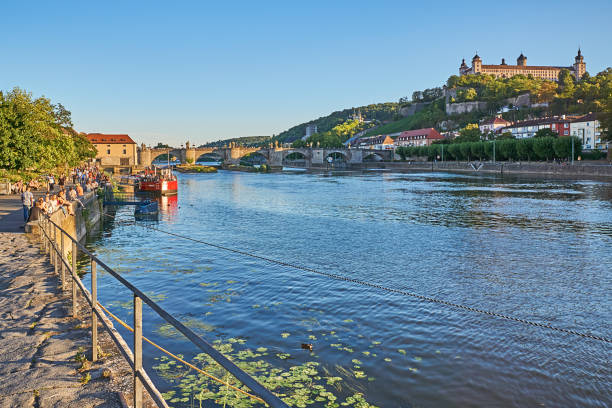 Wuerzburg, Germany - August 06, 2022: People sit on the banks of the Main at sunset in Würzburg. Marienberg Fortress on the right. In the background the old Main Bridge.