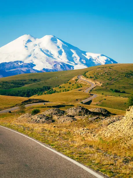A beautiful bend of the road through the green hills of the Caucasus to the main mountain - Elbrus. Elbrus with two peaks against the blue sky is covered with snow. Copy space.