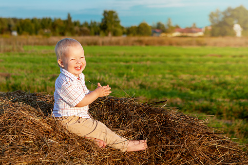 An emotional portrait of a happy little boy sitting on a haystack in a field and enjoying a warm summer day