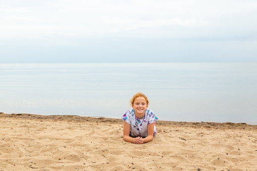 Front view of a young girl laying on her stomach on the beach on a cloudy summer morning. She is smiling at the camera as rain makes its way across the lake towards her.
