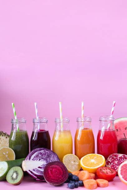 obraz rzędu pięciu szklanych, zakrętkowych butelek zielonych, fioletowych, żółtych, pomarańczowych i czerwonych koktajli z soków owocowych i warzywnych z rozebranymi słomkami do picia, owocami i warzywami, różowym tłem, skupieniem się na pierw - blueberry smoothie glass striped zdjęcia i obrazy z banku zdjęć