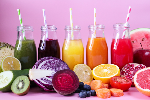Close-up image of row of five glass, screw cap bottles of green, purple, yellow, orange and red fruit and vegetable juice smoothies with stripped drinking straws, fruit and vegetables, pink background, focus on foreground