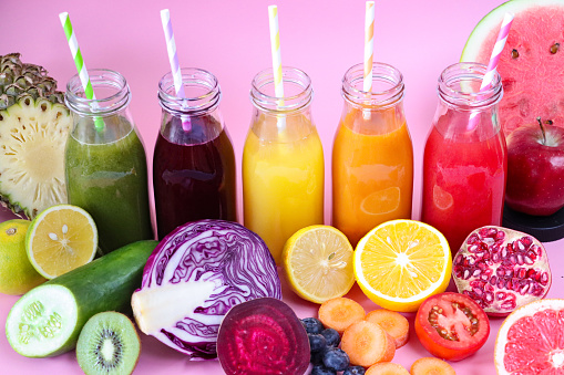 Stock photo showing close-up, elevated view of purple, green, yellow, orange and red fruit and vegetable juice smoothies in glass, screw cap bottles with stripped drinking straws surrounded by watermelon, apple, pomegranate, tomato, carrot, orange, lemon, blueberries, beetroot, red cabbage, kiwi, cucumber, lime and pineapple.