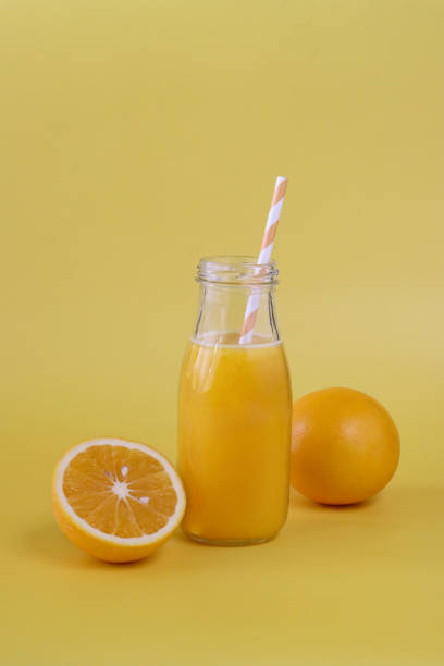 Image of glass, screw cap bottle of orange juice with stripped drinking straw, whole and halved pieces of citrus fruit, yellow background, focus on foreground stock photo