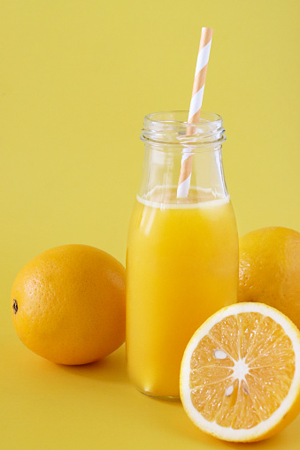 Stock photo showing close-up view of a two, whole pieces of orange fruit and half a citrus fruit displaying the pith surrounding the segmented fruit flesh with seeds, besides a glass, screw cap bottle of orange juice with stripped drinking straw.