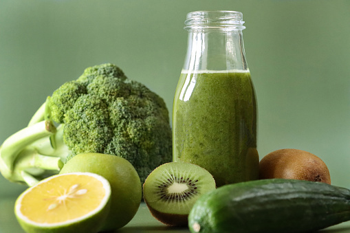 Stock photo showing close-up view of a glass, screw cap bottle of green fruit and vegetable juice smoothie besides whole and halved pieces of kiwi, apple, lime, cucumber and broccoli.