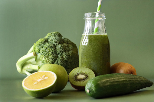 Stock photo showing close-up view of a glass, screw cap bottle of green fruit and vegetable juice smoothie with stripped drinking straw besides whole and halved pieces of kiwi, apple, lime, cucumber and broccoli.