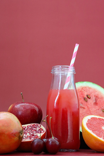 Stock photo showing close-up view of a glass, screw cap bottle of red fruit juice smoothie with stripped drinking straw besides whole and halved pieces of pomegranate, apple, pear, cherries, tomato, watermelon and pink grapefruit.