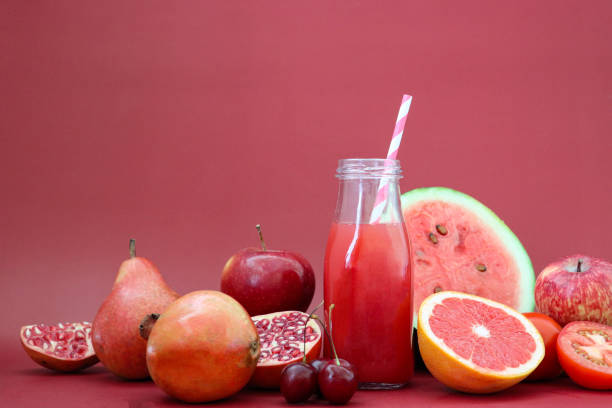 Close-up image of glass, screw cap bottle of red fruit juice smoothie with stripped drinking straw, pomegranate, apple, pear, cherries, tomato, watermelon and pink grapefruit, whole and halved pieces of fruit, red background, copy space stock photo