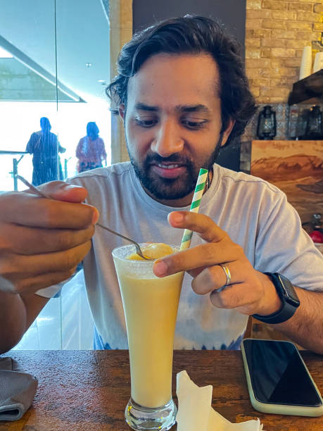 istock Image of Indian man using long handled spoon to eat ice cream from vanilla milkshake drink, tall glass with stripped green and white paper drinking straw, restaurant setting, focus on foreground 1414024645