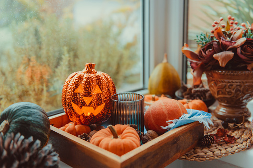 Halloween cozy mood composition on the windowsill. Lighting jack-o-lantern, decorative pumpkins, cones, candles on wooden tray. Hygge Halloween home decor. Selective focus