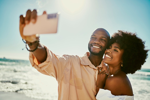 Happy, trendy and young couple taking a selfie while having fun at the beach together. Carefree tourists enjoying their seaside vacation, celebrating their freedom and time together outdoors