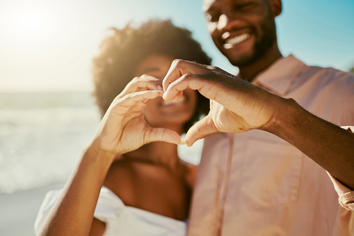 A loving couple making heart gesture outdoors on a date at the beach on a sunny summer day. Closeup portrait of African American lovers showing love together while enjoying the weekend