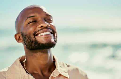 Happy African man smiling while standing at the beach enjoying relaxing and warm sunlight on hot day in summer. Close up portrait of handsome, free and cheerful male with the sea in the background