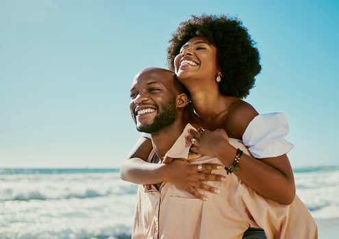 Happy, carefree and in love couple at the beach enjoying a relaxing summer vacation outdoors with sea waves and blue sky copy space background. Young, loving and playful man giving woman a piggyback