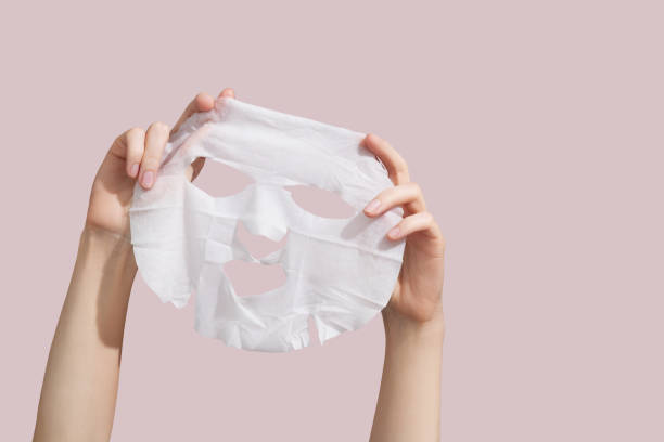 Female hands holding sheet of white mask on pink background. stock photo
