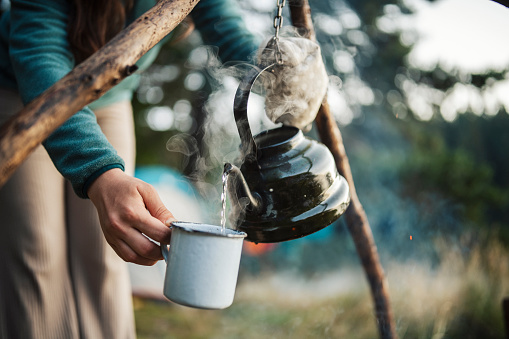 Close up shot of unrecognizable woman pouring hot tea in mug from retro kettle over campfire in the morning.