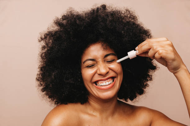 Beautiful young woman applying cosmetic oil on her face Beautiful young woman smiling cheerfully while applying face serum using a dropper. Happy woman with Afro hair treating her skin with a nourishing beauty product. grooming product photos stock pictures, royalty-free photos & images