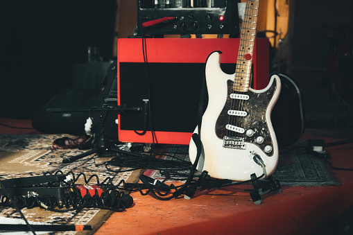 A white electric guitar and a red amplifier onstage with electrical audio cables and sound effects foot pedals