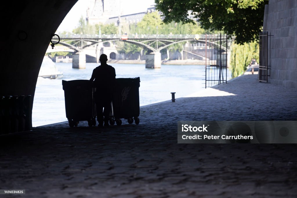Man in silhouette pushing garbage bins under an arch by the Seine River Under Pont Neuf on the south bank of Île de la Cite, Paris Paris - France Stock Photo