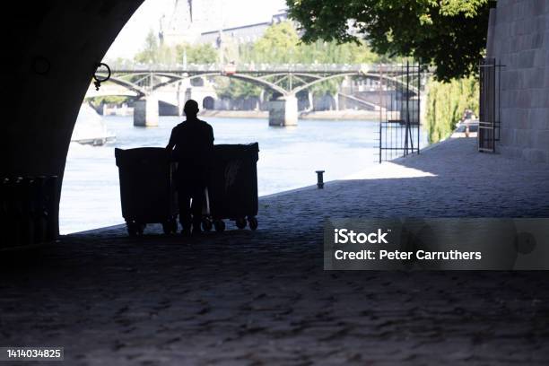 Man In Silhouette Pushing Garbage Bins Under An Arch By The Seine River Stock Photo - Download Image Now