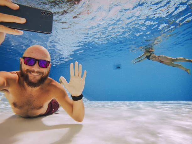 Selfie with mobile phone underwater: videocall with friends Funny man selfie with mobile phone underwater: woman swimming in the background photo bomb stock pictures, royalty-free photos & images