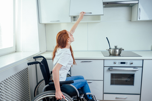 disabled redhaired ginger woman in wheelchair preparing meal in kitchen ,try to reaching the top shelf.