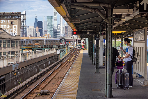 Queensboro Plaza Station, Long Island City, Queens, New York, NY, USA - July 2 2022: Young woman checking her phone while the train is approaching the elevated subway station in front of the Manhattan skyline