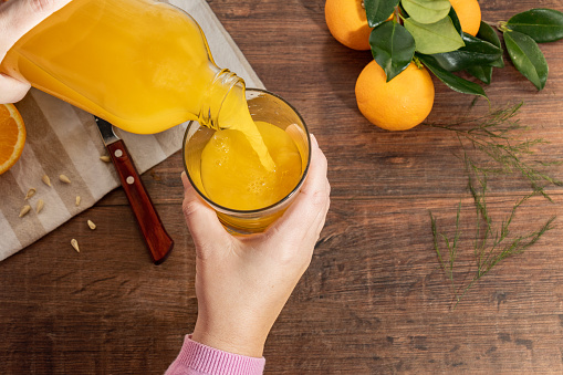 a person pouring orange juice into a glass cup. in the background you can see whole oranges and a knife resting on a wooden table