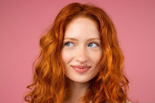 Close up photo of a beautiful redhead lady with blue attractive eyes looking aside over a pink background with smiling facial expression. High quality photo