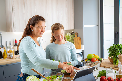 Happy mother and her daughter preparing healthy vegetables salad in kitchen. They are using a digital tablet to get healhy vegetables recipe. High resolution 42Mp indoors digital capture taken with SONY A7rII and Zeiss Batis 40mm F2.0 CF lens