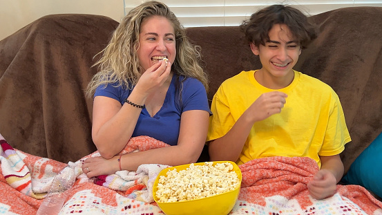 Latino mother and her teenage son are eating popcorn while they watch tv together. They are sitting on an old beat up couch that they cannot afford to replace. He has braces on his teeth that were paid for with government aid.