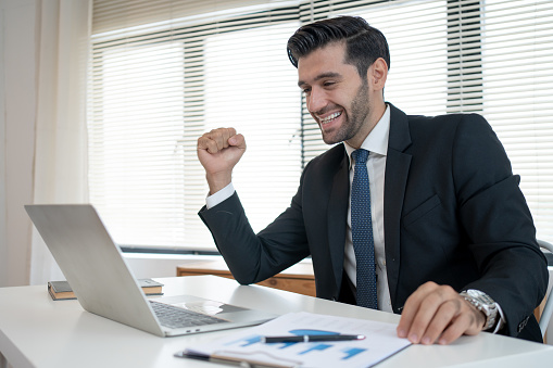 Businessman raising his fist, rejoicing at the success of the financial investment according to the laptop earnings report.